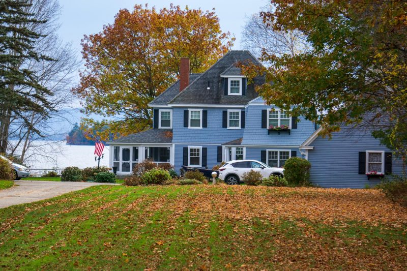 A blue house surrounded by vibrant autumn leaves near a calm lake in a rural setting.