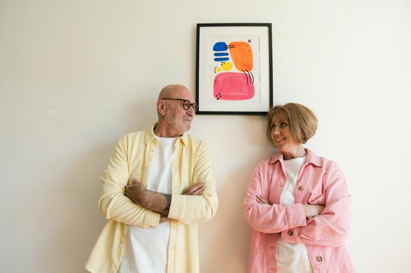 Elderly couple smiling at each other under colorful artwork in a cozy indoor setting.
