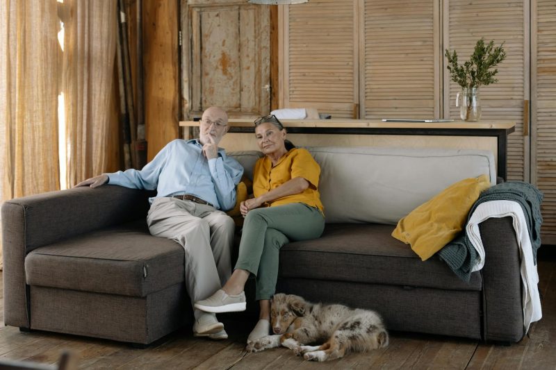 Elderly couple cuddles on a sofa with their dog in a cozy, rustic living room.