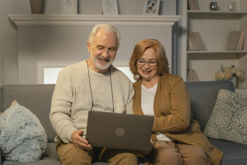 Elderly couple smiling together on a cozy couch while using a laptop.