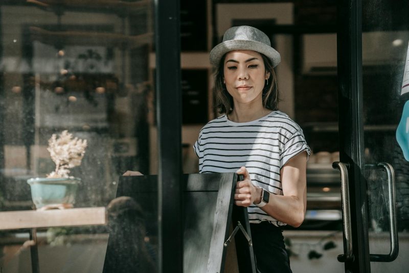 Woman in casual wear and hat placing a sign outside a modern cafe on a sunny day.