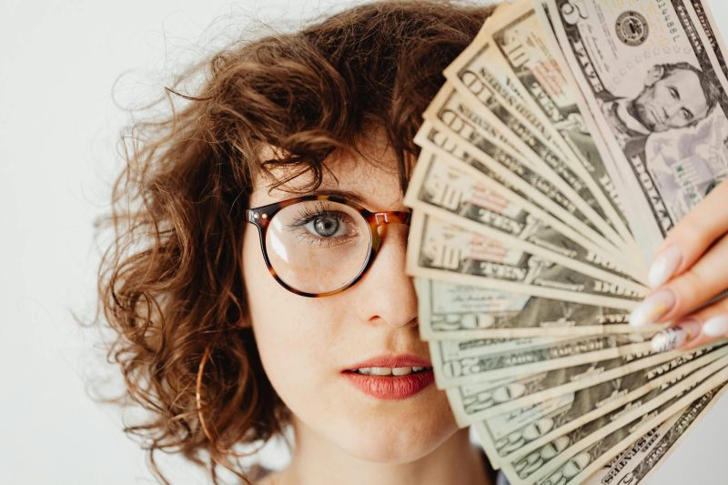 Caucasian woman with eyeglasses holding several US dollar bills, smiling confidently.