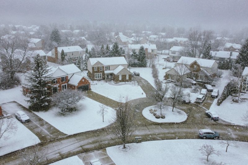 Aerial view of a snowy suburban neighborhood with houses, tree-lined streets, and quiet, empty sidewalks.