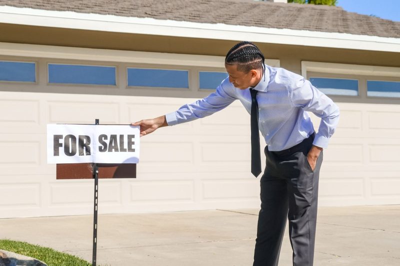 Real estate agent placing a "For Sale" sign outside a suburban house on a sunny day.